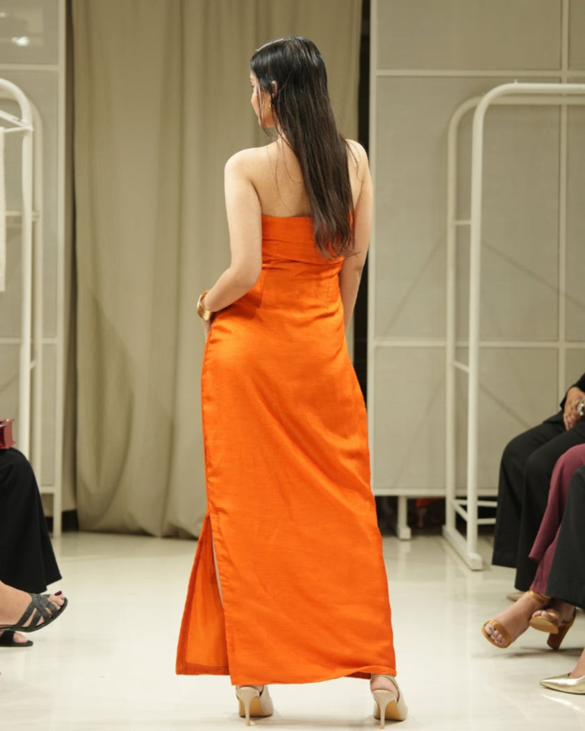 Model in an orange dress walking on a catwalk with an audience taking photos.