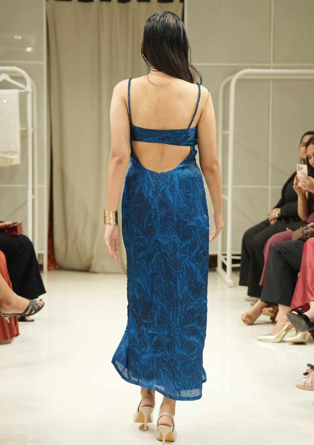 Model wearing a blue lace dress walking on a catwalk with an audience seated on either side.