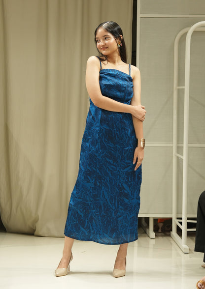 Woman wearing a blue dress standing in a room with beige curtains and a white shelf.